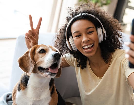 Happy young african woman in headphones relaxing on a couch with her pet dog at home, taing a selfie