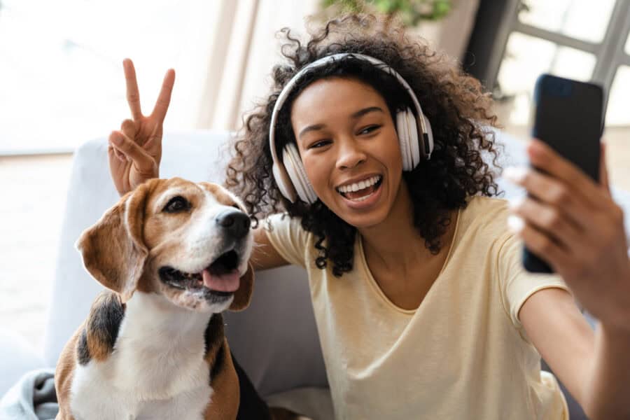 Happy young african woman in headphones relaxing on a couch with her pet dog at home, taing a selfie