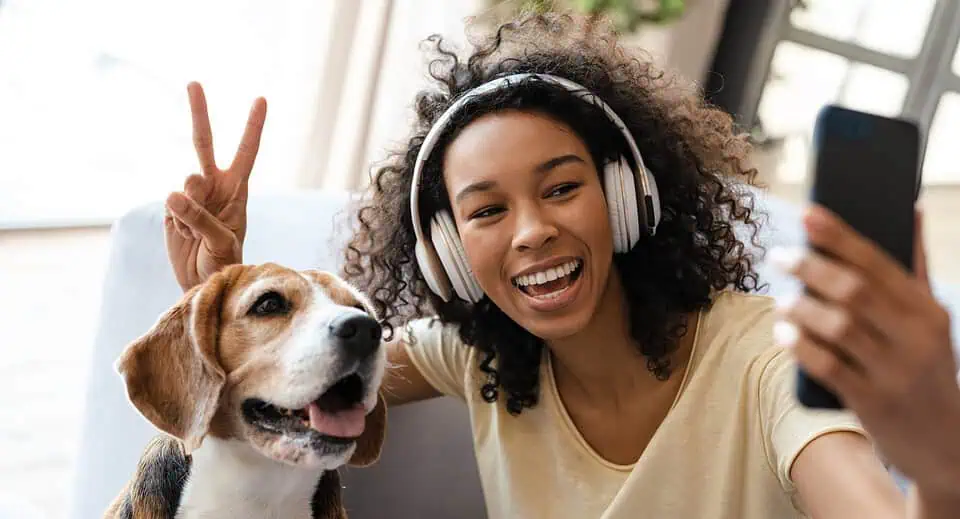 Happy young african woman in headphones relaxing on a couch with her pet dog at home, taing a selfie