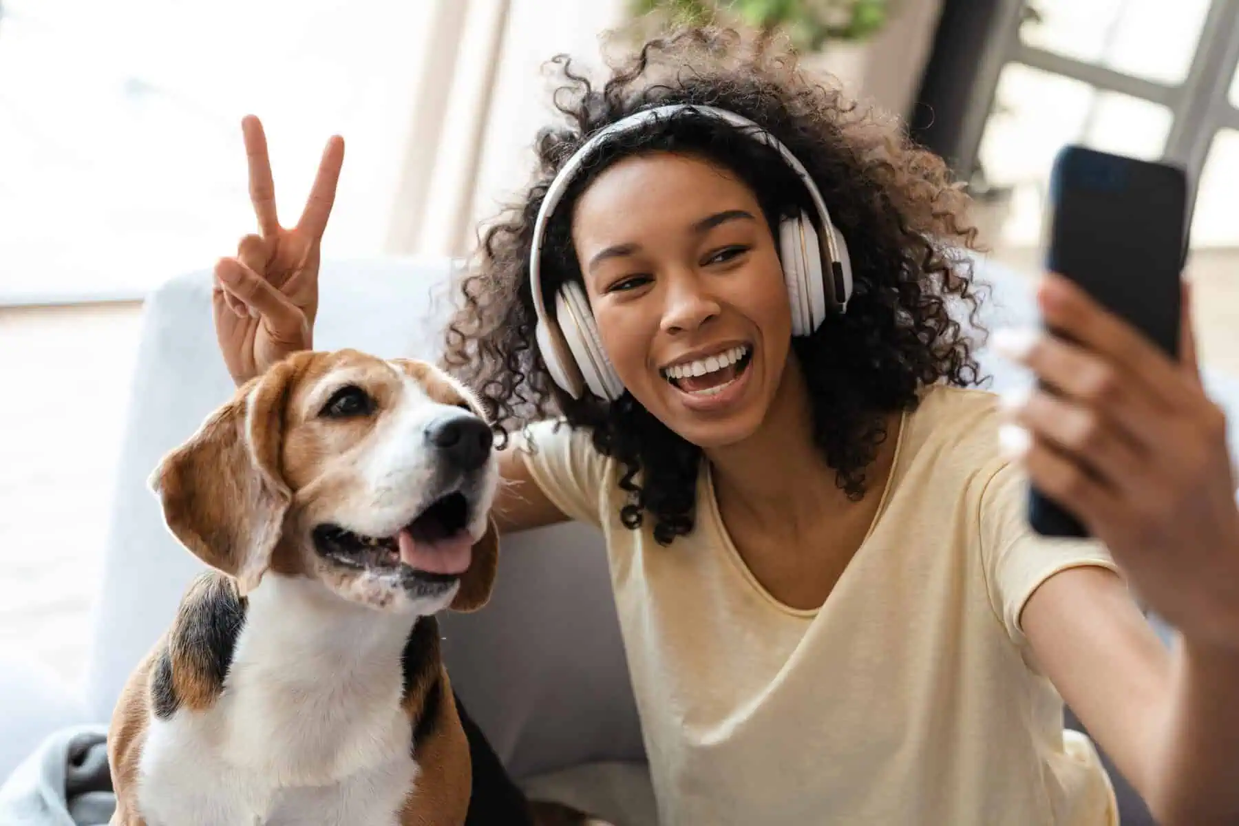Happy young african woman in headphones relaxing on a couch with her pet dog at home, taing a selfie